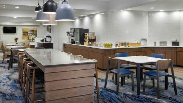A modern hotel dining area with a long counter, multiple seating options, and a kitchen in the background. - Fairfield Inn & Suites Jacksonville Airport (Jacksonville)