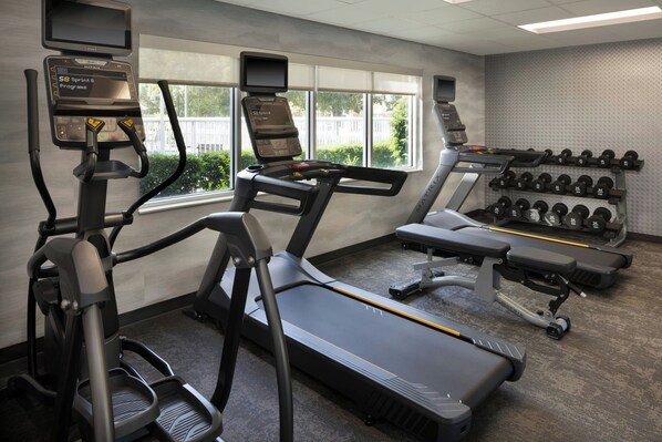 Un gimnasio con cintas de correr, una elíptica y un banco para press de banca. Hay mancuernas en un estante y un televisor montado en la pared.