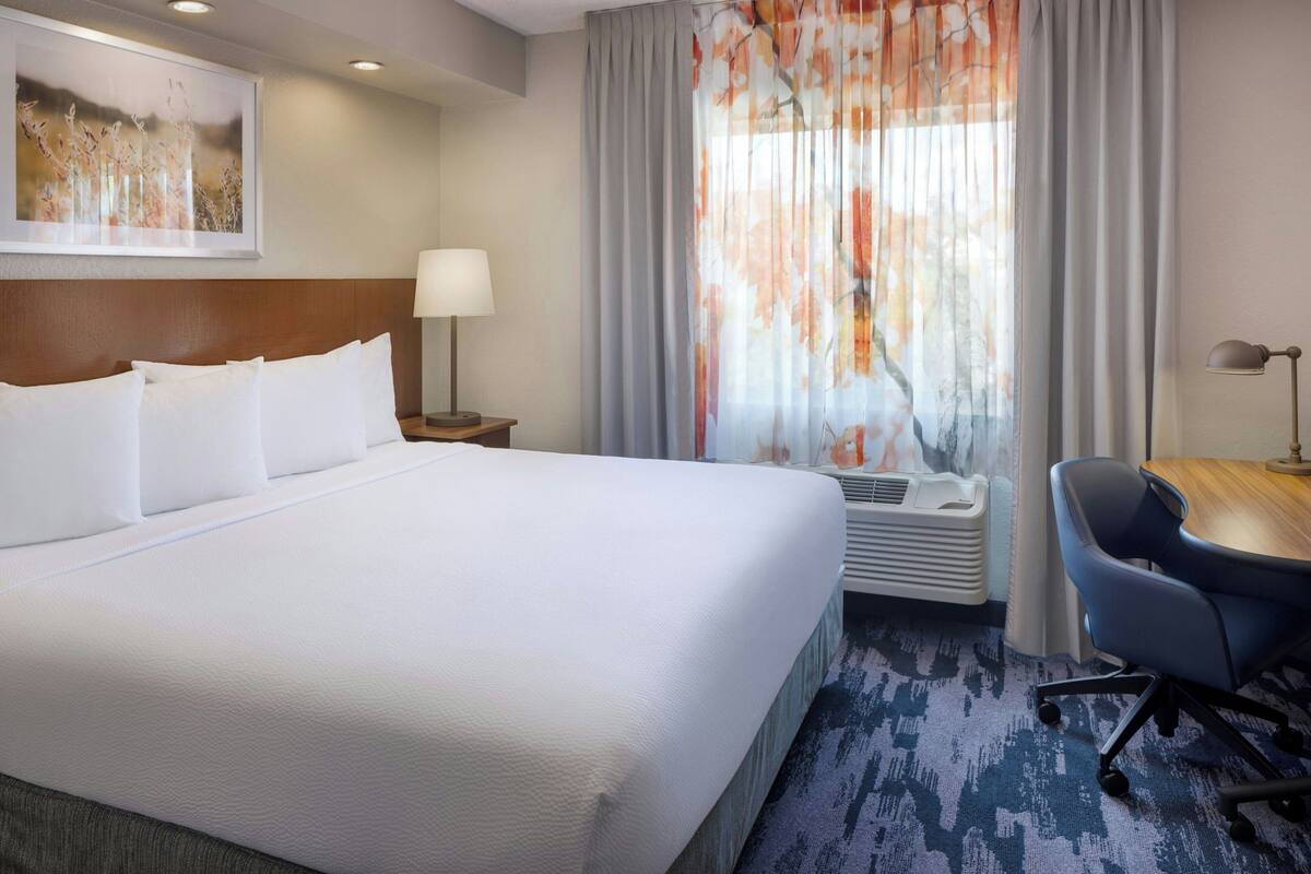 A hotel room with a large bed, a desk, a chair, a lamp, and a window with curtains.