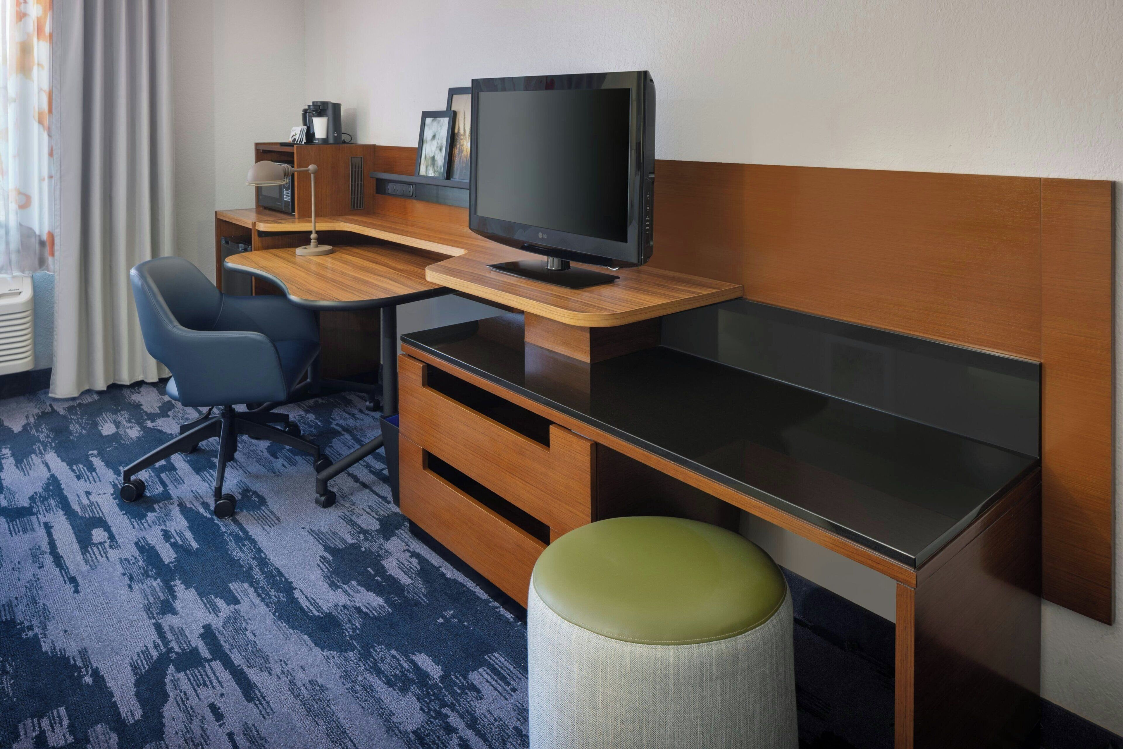 Habitación de hotel con un escritorio de madera, una computadora, un televisor, una silla y un taburete pequeño.