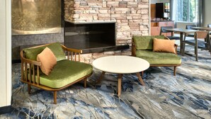 Una habitación con chimenea de piedra, dos sillones tapizados en verde, una mesa redonda y un gran ventanal con cortinas.