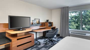 A hotel room with a bed, a desk with a flat-screen TV, a chair, a microwave, and a window with a view of trees.