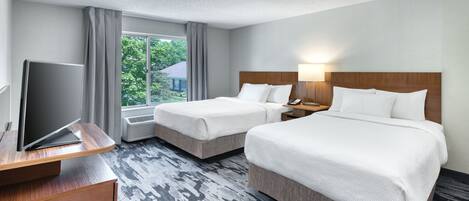 A hotel room with two beds, a desk with a flat-screen TV, a nightstand, and a window with a view of greenery.