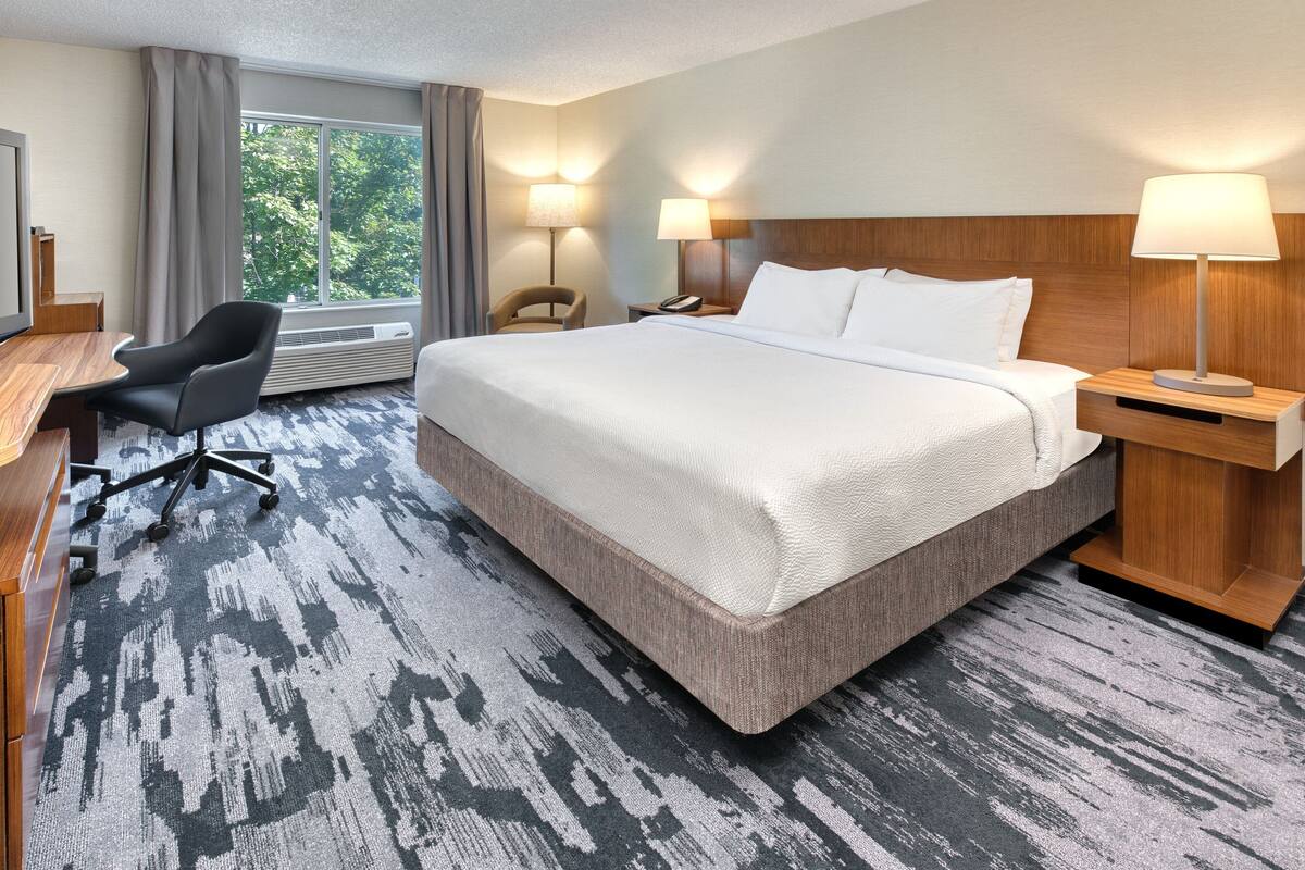 A hotel room with a large bed, a desk with a chair, two bedside lamps, and a view of trees through the window.