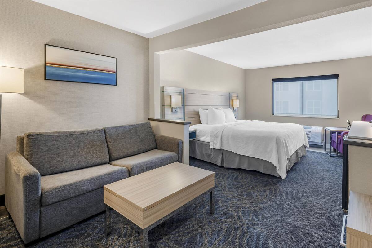 Ein Hotelzimmer mit grauem Sofa, einem Holztisch, einem Bett mit weißen Leinen, einem Kopfteil mit wandmontierten Lampen und einem großen Fenster.