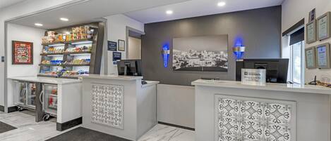 Un área de recepción con mostrador, pantalla digital, cuadro con vista urbana y refrigerador con bebidas.