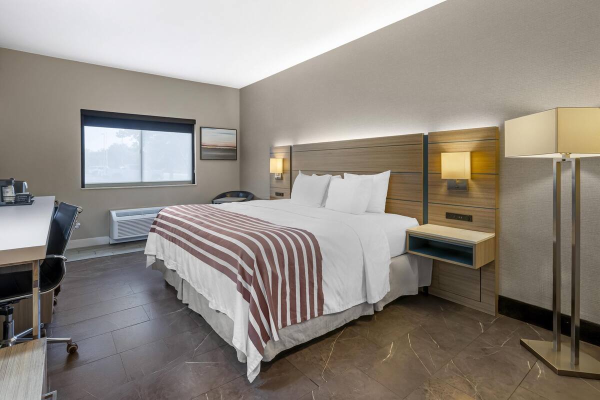 Ein Hotelzimmer mit einem großen Bett, einem Schreibtisch und einem Sessel. Ein Fenster bietet einen Ausblick nach draußen.