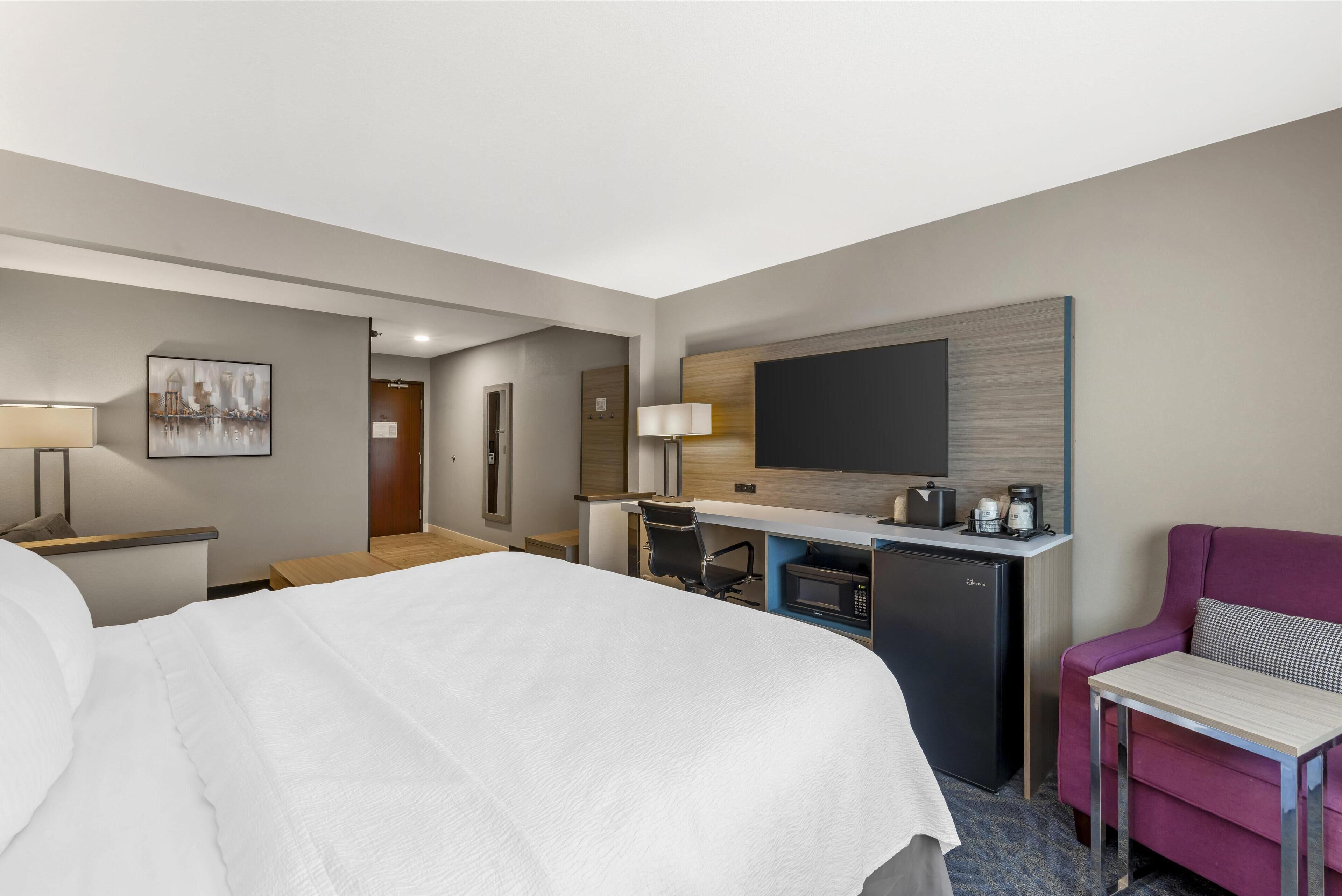 A hotel room with a bed, a desk with a computer, a microwave, a coffee maker, a TV, and a chair.