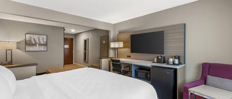 A hotel room with a bed, a desk with a computer, a microwave, a coffee maker, a TV, and a chair.