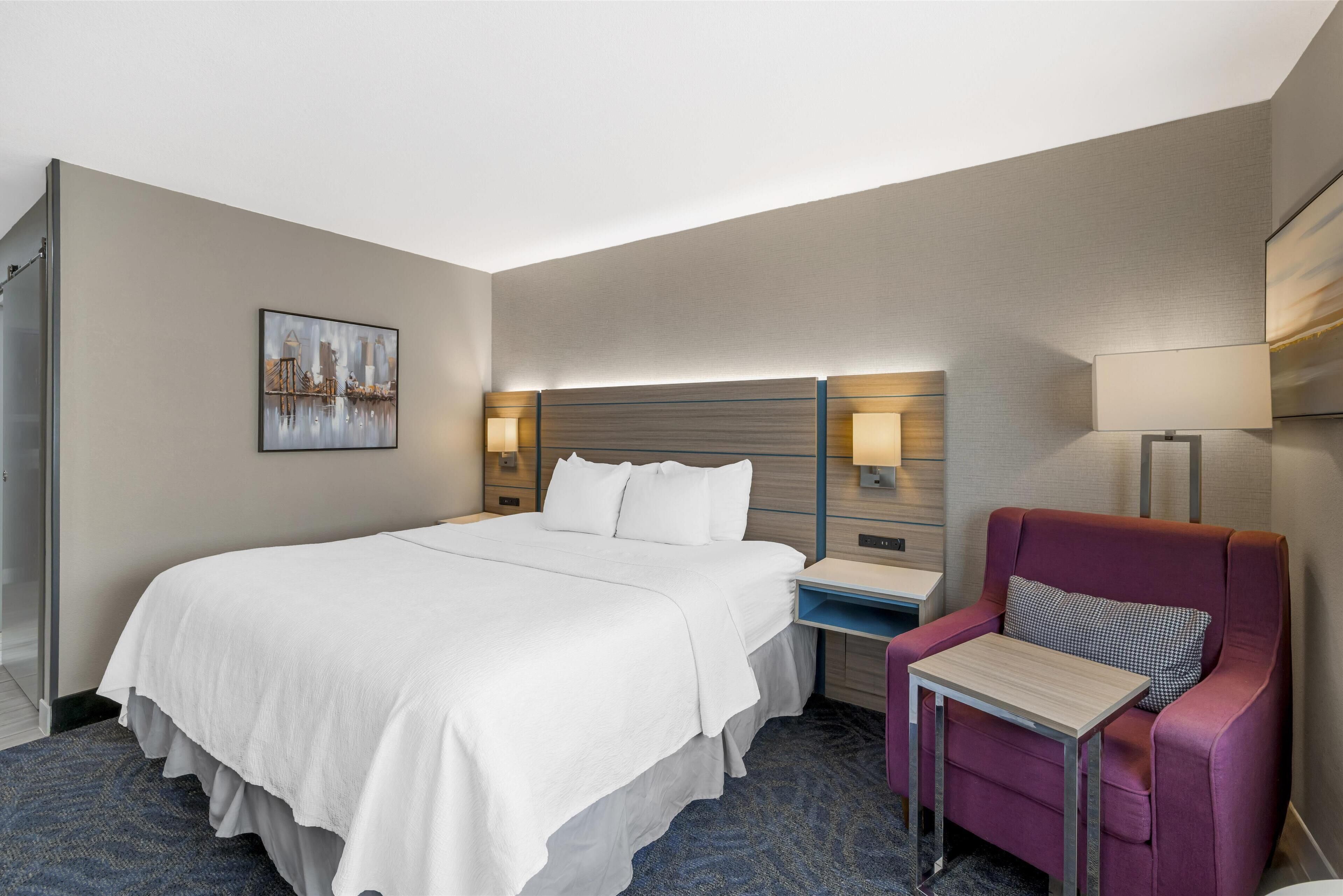 Habitación de hotel con una cama grande, una mesita de noche con lámpara, una silla y un cuadro en la pared.