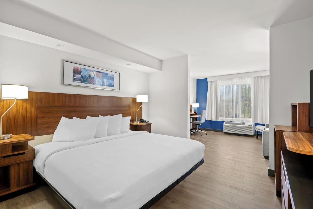 A hotel room with a large bed, wooden headboard, bedside tables with lamps, a desk with a chair, and a large window with curtains.