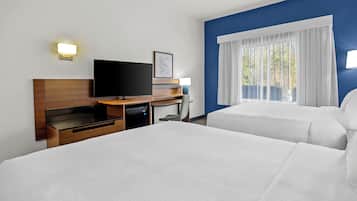 Habitación de hotel con dos camas, un televisor de pantalla plana, un escritorio con lámpara y una ventana con cortinas blancas.