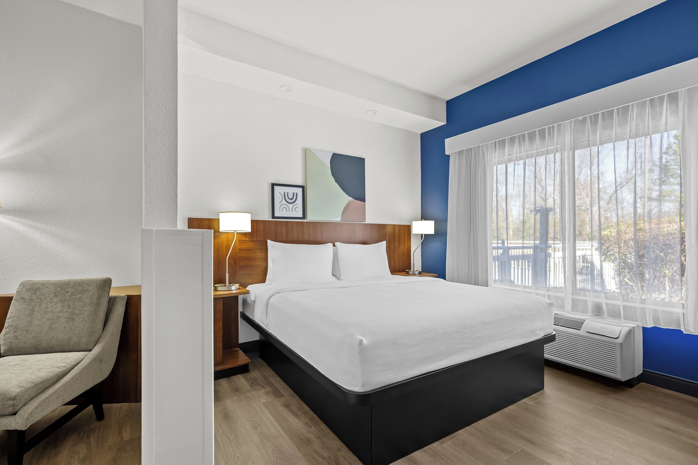 Une chambre d’hôtel avec un grand lit, une chaise, une table de chevet avec une lampe, une fenêtre avec des rideaux et un climatiseur fixé au mur.
