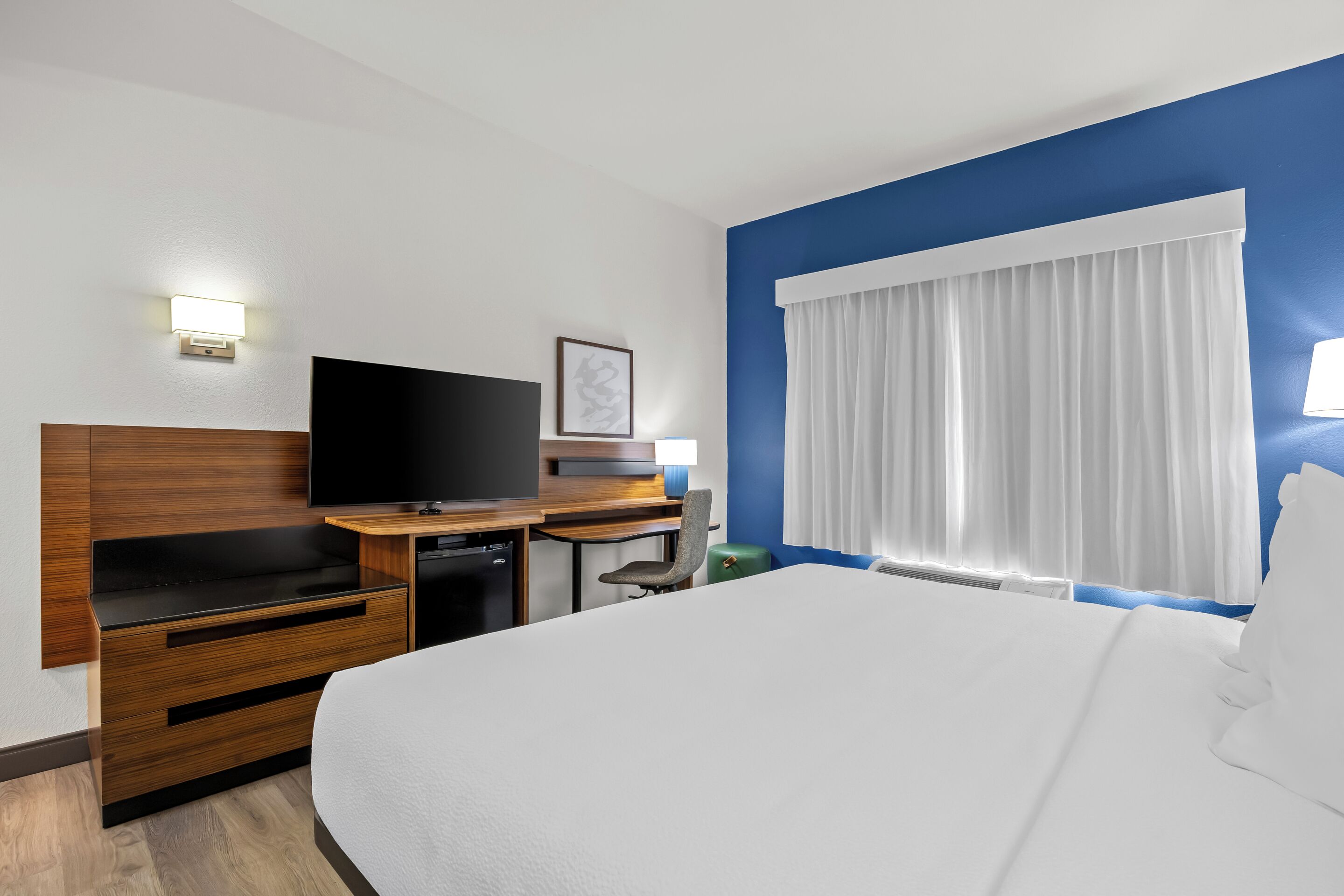 A hotel room with a large bed, a flat-screen TV, a desk with a computer, a chair, and a window with white curtains.