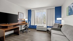 A hotel room with a wooden desk, a grey sofa, a TV, a chair, a window with curtains, and a wall-mounted air conditioner. - Spark by Hilton Macon North (Macon)