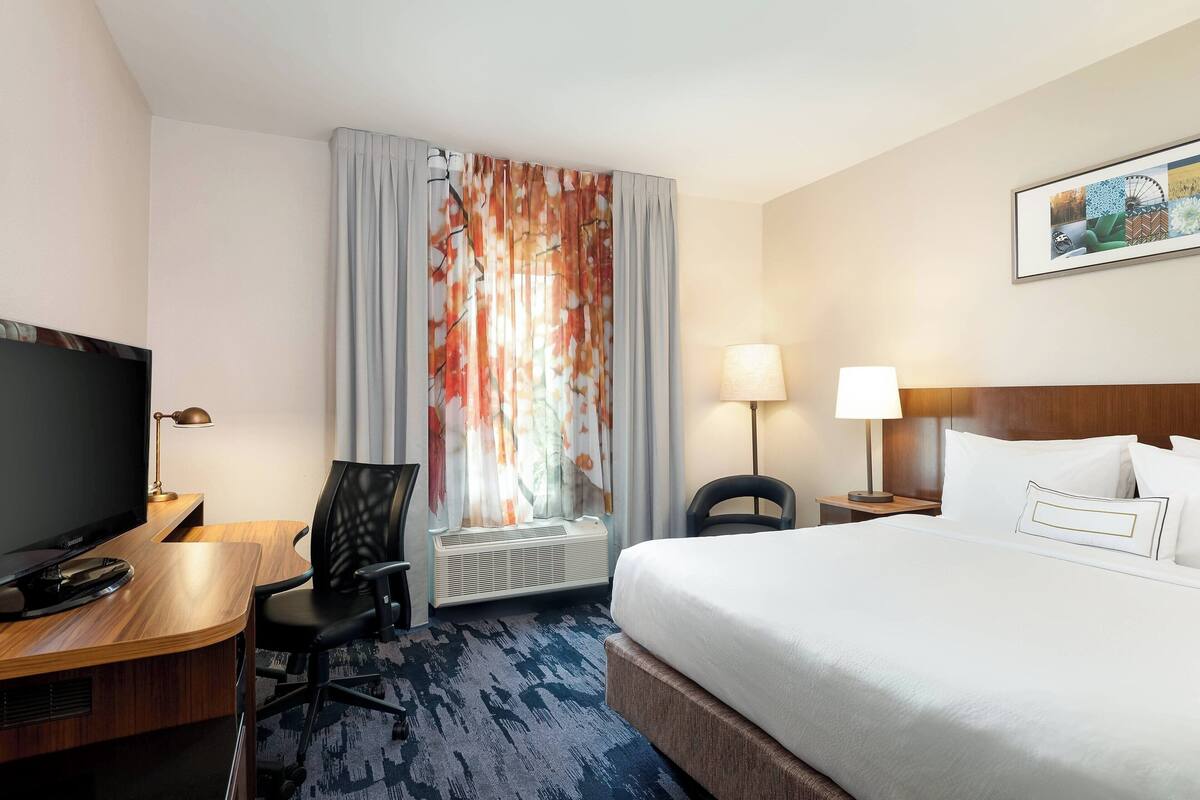 Une chambre d’hôtel comprenant un lit, un bureau avec un ordinateur, une télévision, une chaise, une lampe et une fenêtre avec des rideaux.