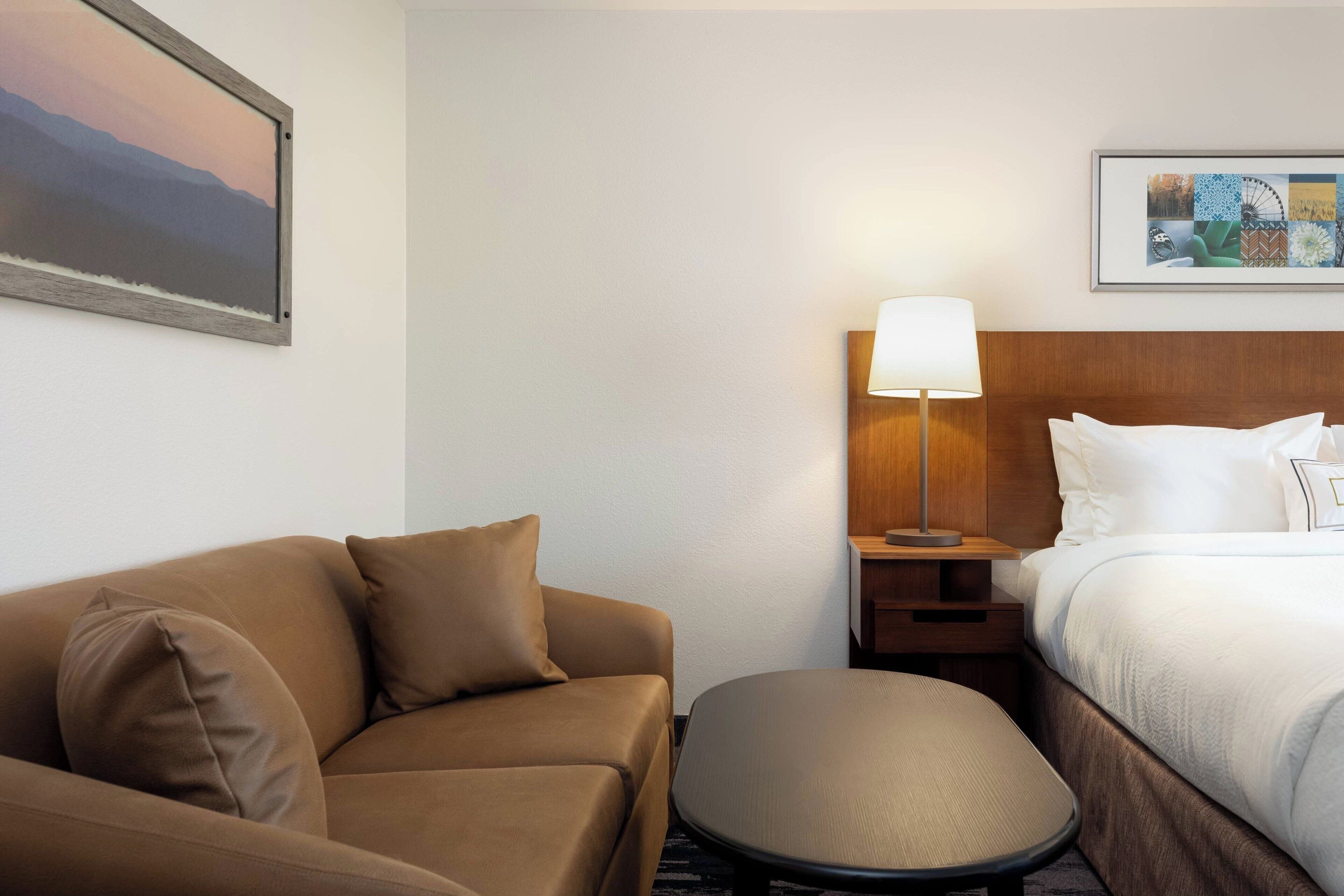 Habitación de hotel con cama, sofá, sillón, mesita de noche con lámpara y dos cuadros en las paredes.