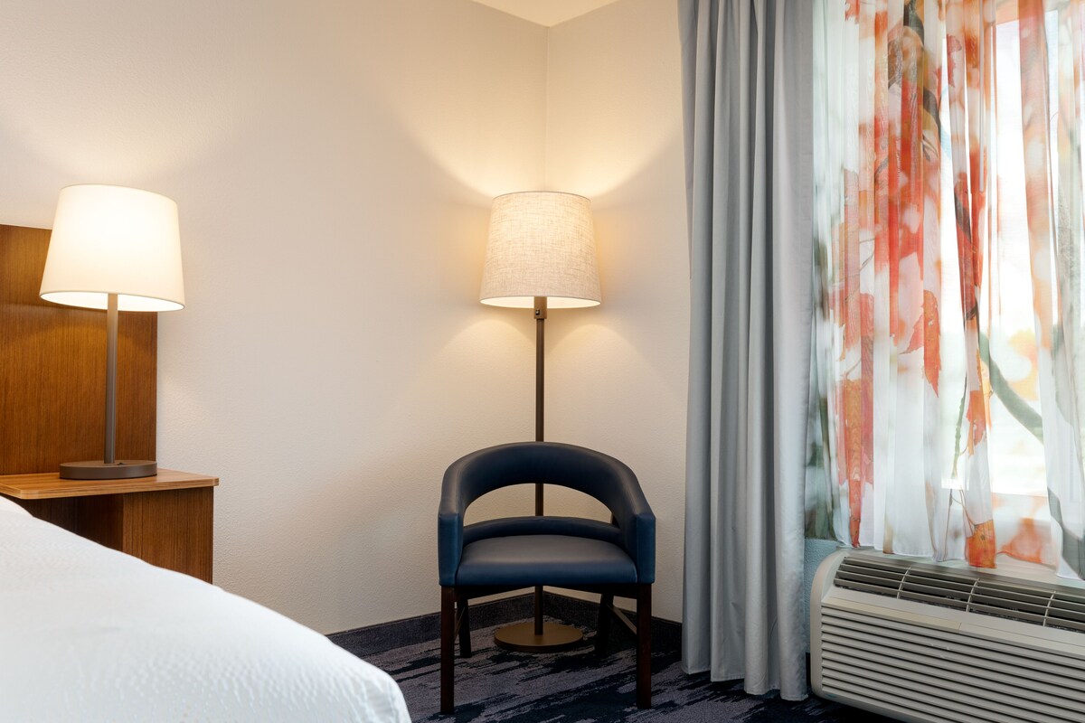 Ein Hotelzimmer mit einem Bett, einem Schreibtisch mit Lampe, einem Sessel und einem Fenster mit Vorhängen.