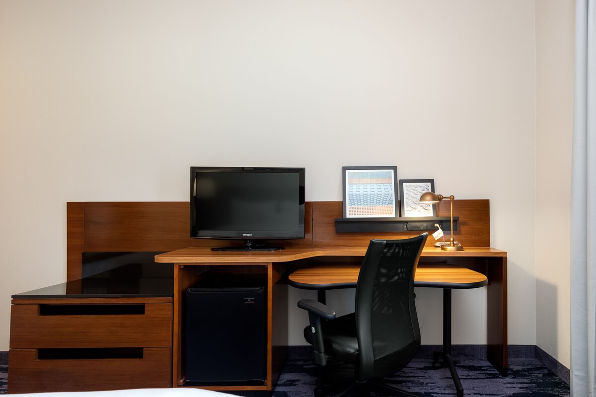 Een hotelkamer met een houten bureau, een televisie, een kleine koelkast, een stoel en een bureaulamp.