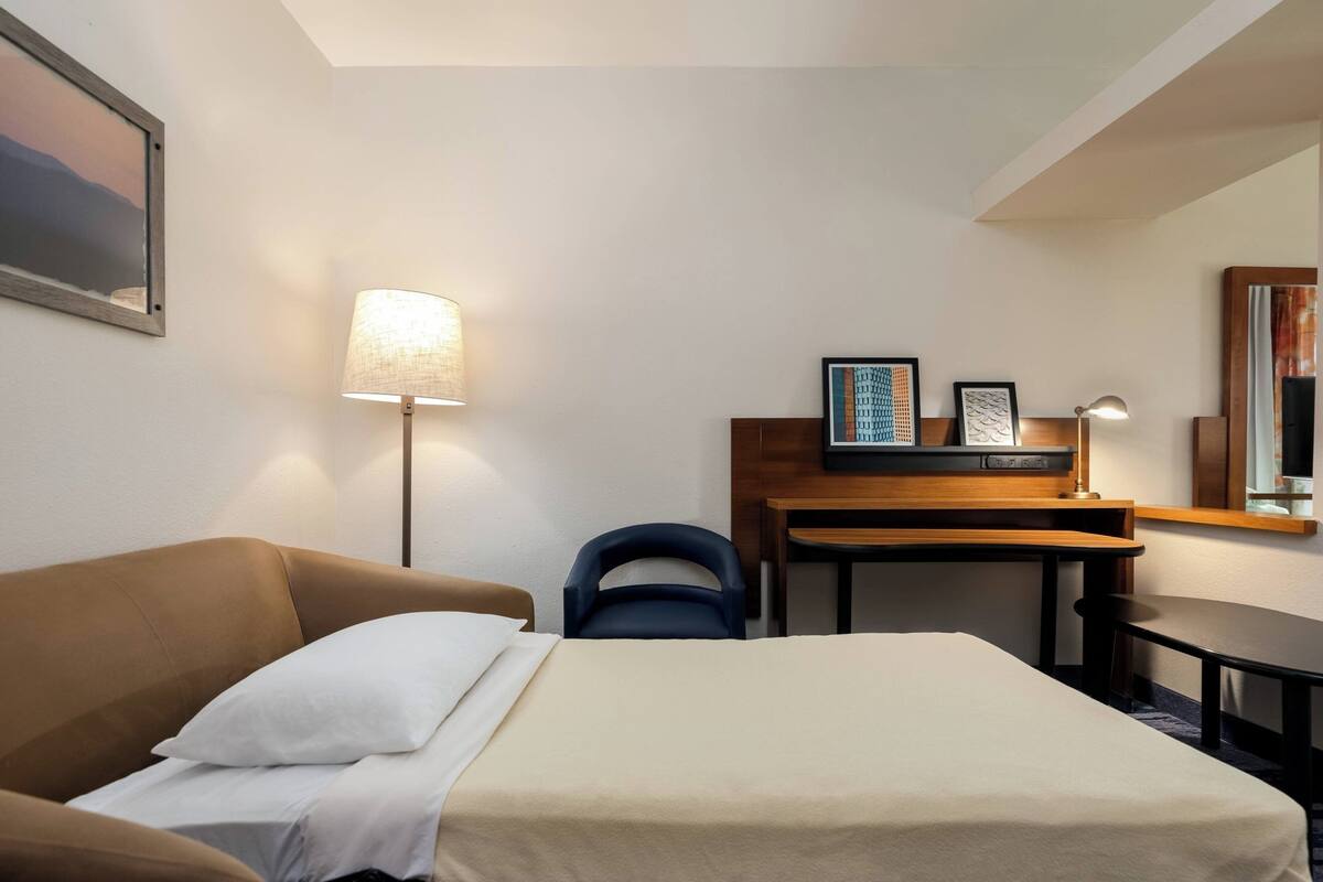 Une chambre d’hôtel avec un lit, un bureau, une chaise, une lampe et un tableau au mur.