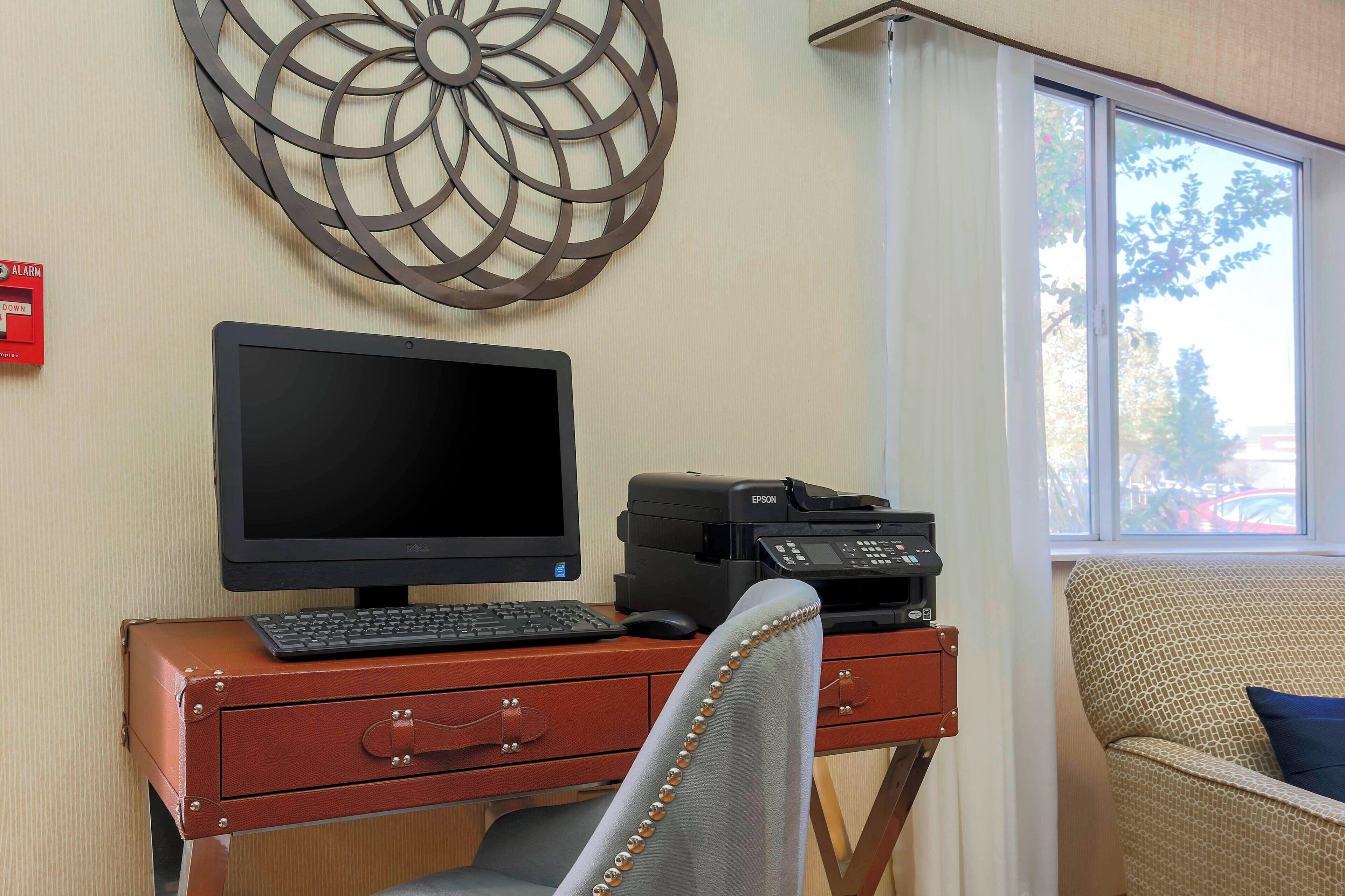 A workspace with a monitor, keyboard, and printer on a desk, a chair with a decorative backrest, and a wall-mounted decorative piece.