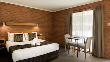 Habitación de hotel con pared de ladrillo, cama con almohadas estampadas, mesita de noche con lámpara, una pequeña mesa con sillas y ventana con cortinas.