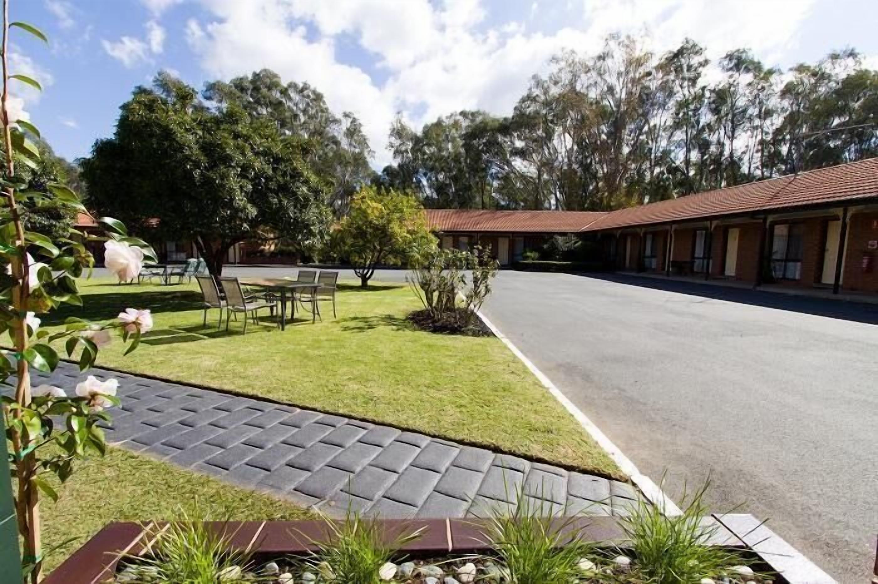 A motel with a paved driveway, a lawn with a table and chairs, and a row of rooms with a red roof.