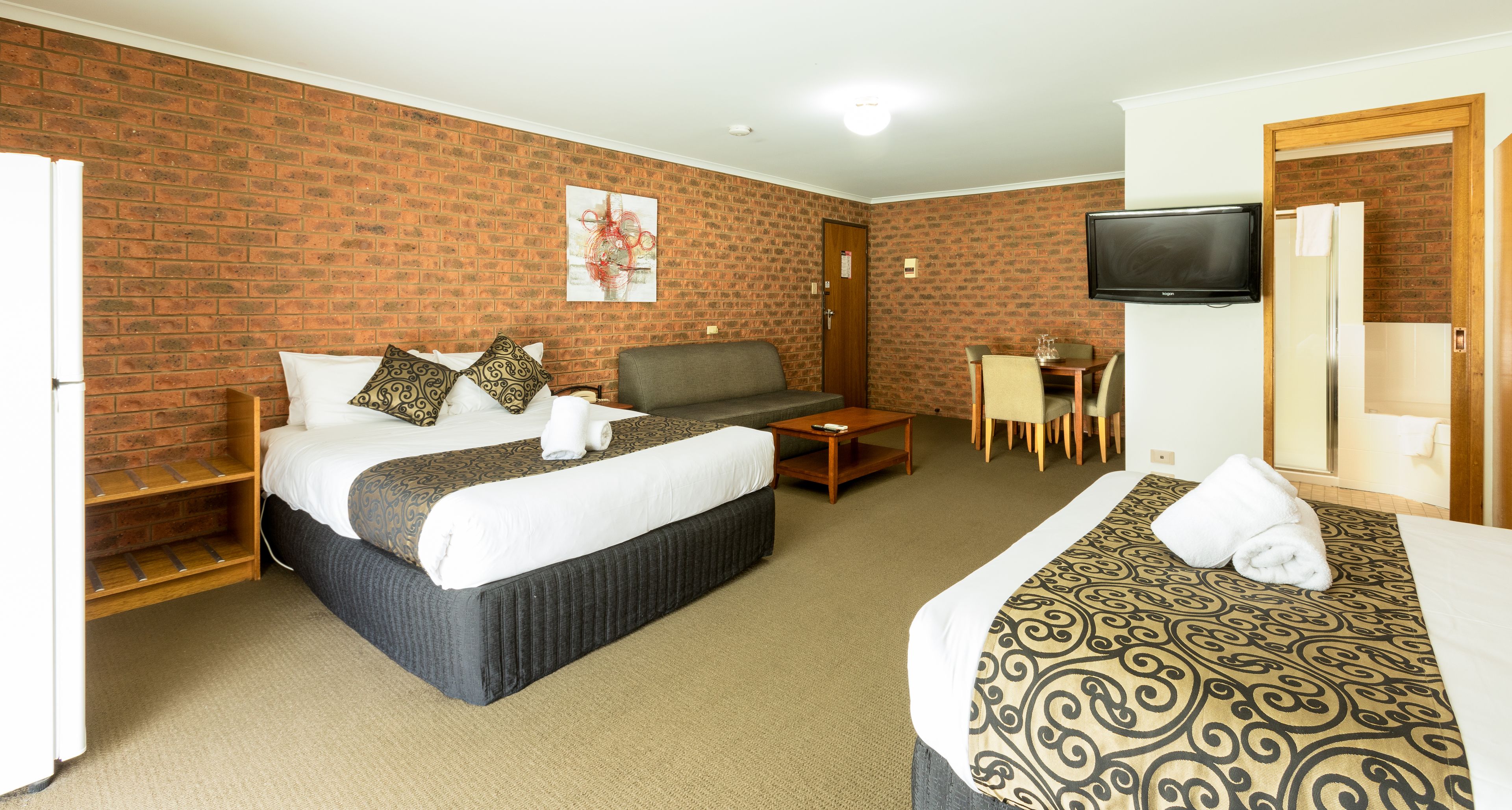 A hotel room with a brick wall, a bed with patterned bedding, a sofa, a small table, a TV, and a shower area.