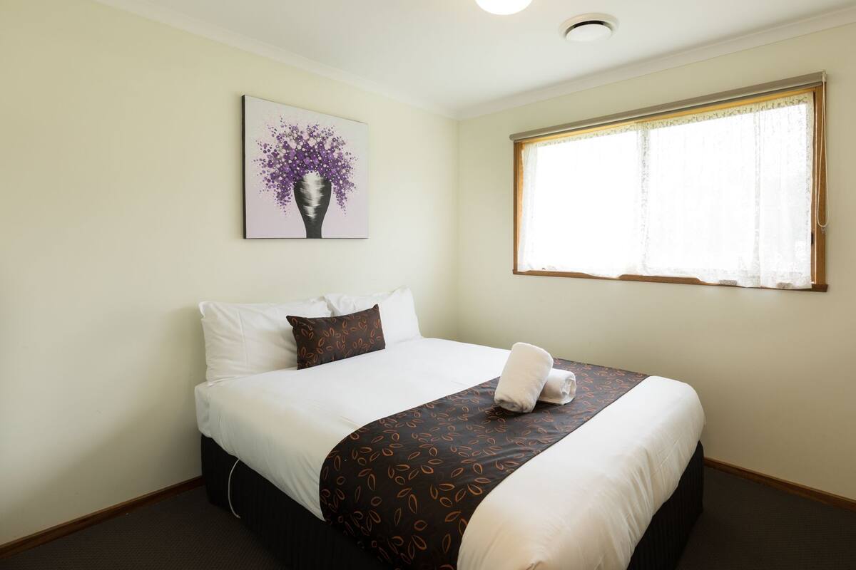 Una cama bien tendida con una colcha estampada, un almohadón y un cojín decorativo en la cabecera. Un cuadro enmarcado con flores moradas cuelga en la pared sobre la cama.