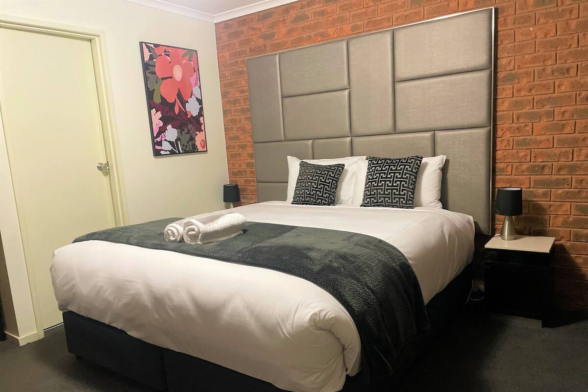 Una cama bien hecha con un cabecero gris, una toalla blanca enrollada sobre la cama y una almohada con estampado blanco y negro.