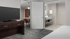 A hotel room with a bed, a TV on a wooden dresser, a bathroom with a mirror and towel rack, and a patterned rug.