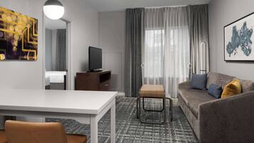 A modern hotel room with a white desk, a sofa with cushions, a television, a window with curtains, and artwork on the walls.