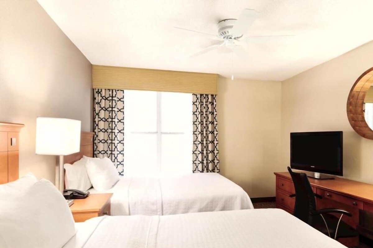 Une chambre d’hôtel avec deux lits, un bureau avec une télévision, une lampe et une fenêtre avec des rideaux à motifs.