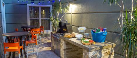 Un patio exterior con una estación de barbacoa de piedra, sillas naranjas y una mesa de comedor.
