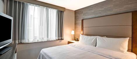 Camera d'hotel con un letto grande, una TV a schermo piatto, un comodino con lampada e una finestra con tende.