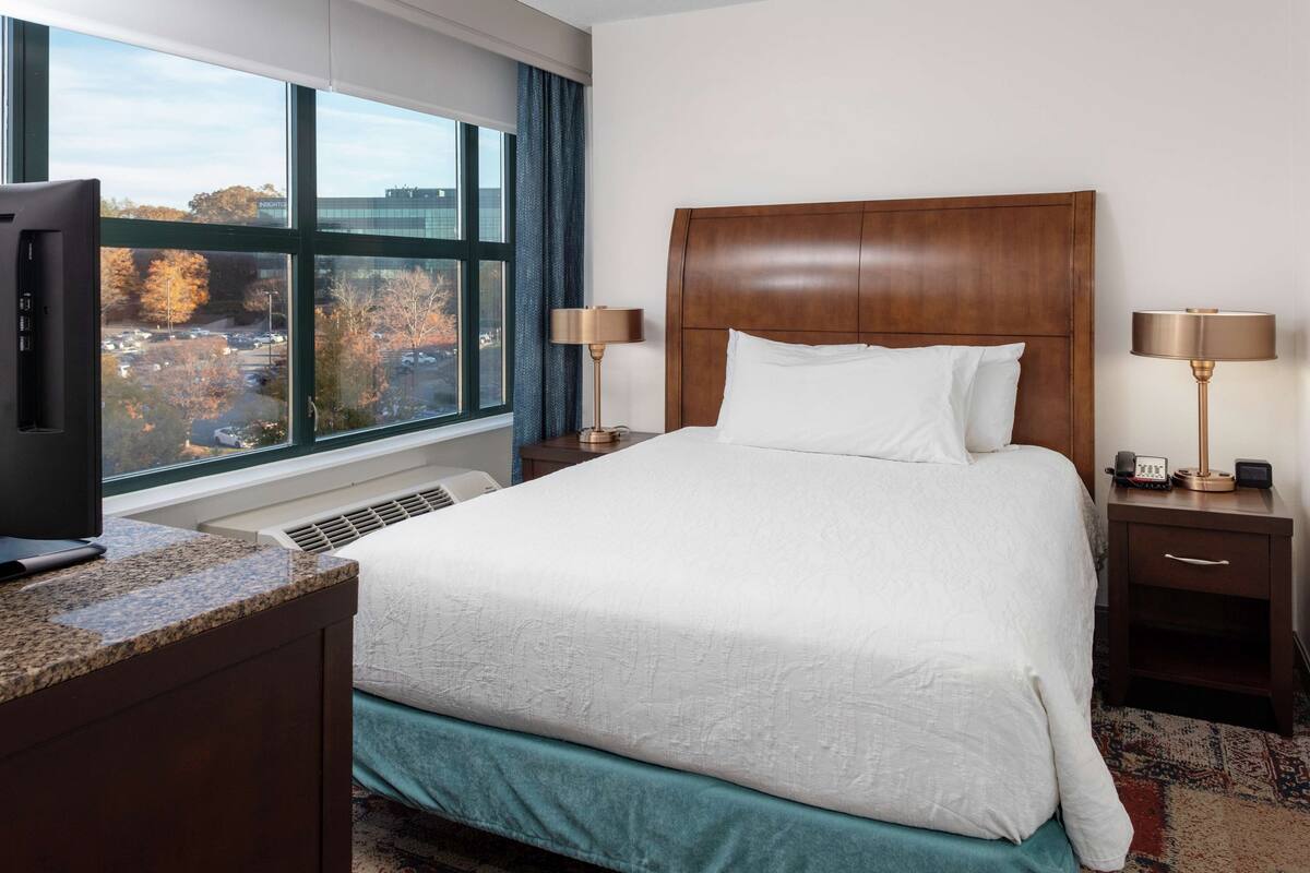 A hotel room with a large bed, a TV on a desk, and a view of a parking lot and trees through the window.