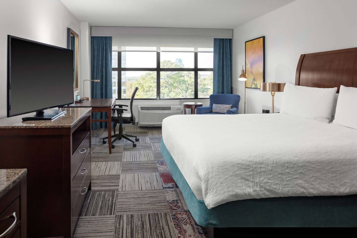 A hotel room with a large bed, a desk with a flat-screen TV, a chair, and a window with a view of trees.