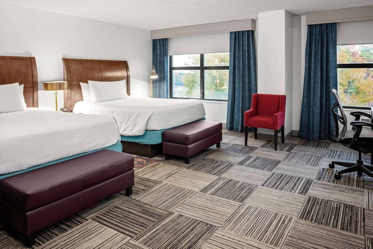 A hotel room with two beds, a red chair, a desk, and a view of trees through the window.