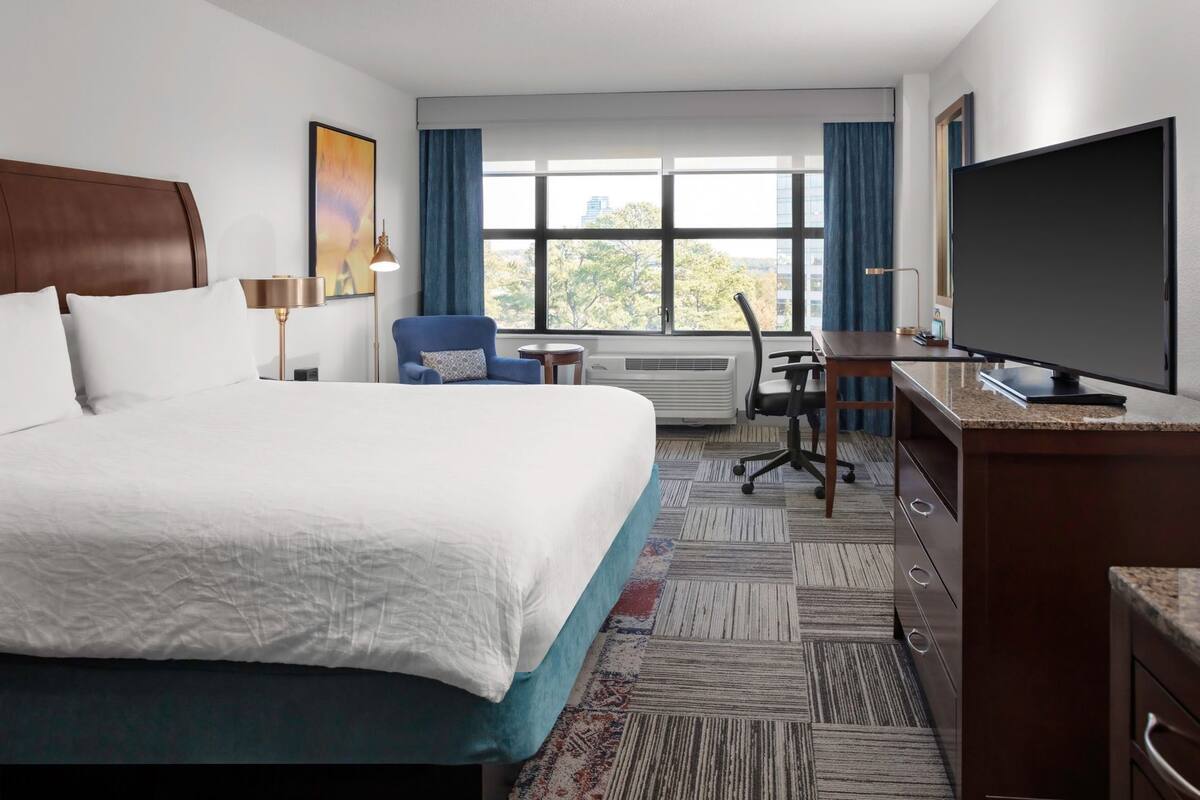 A hotel room with a large bed, a desk, a chair, a flat-screen TV, and a window with curtains.