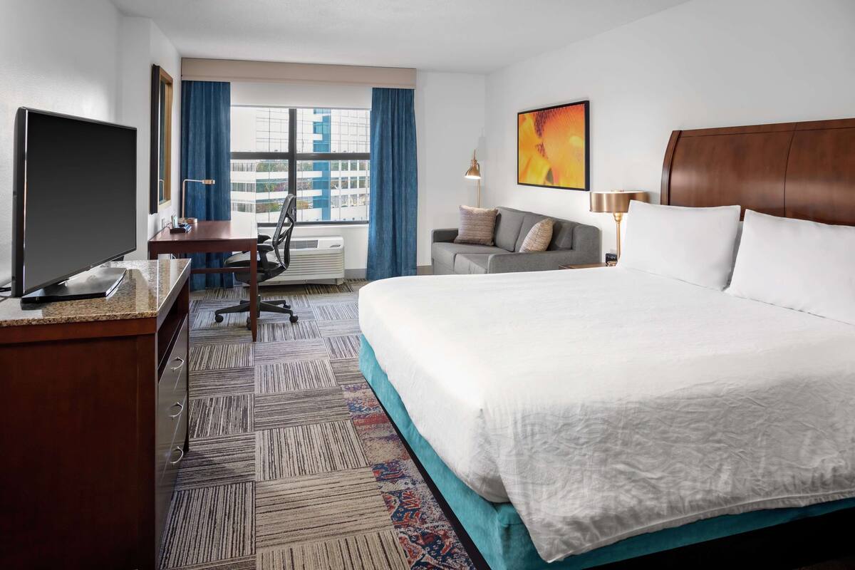 A hotel room with a large bed, a desk with a computer, a chair, a sofa, a TV, and a window with curtains.