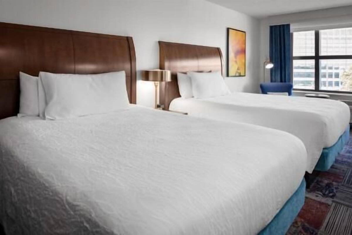 A hotel room with two large beds, a nightstand, a lamp, and a window with a view of buildings.