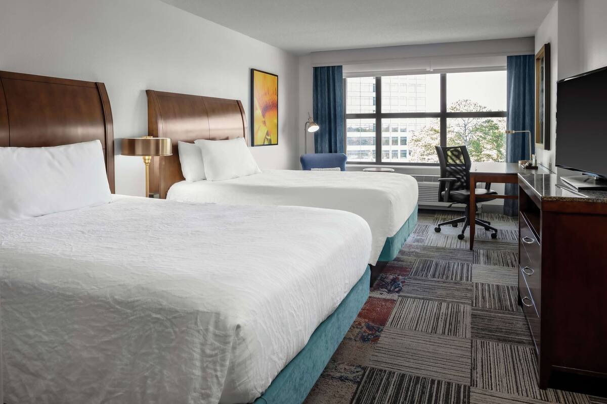 A hotel room with two beds, a desk, a chair, a TV, and a large window with curtains.