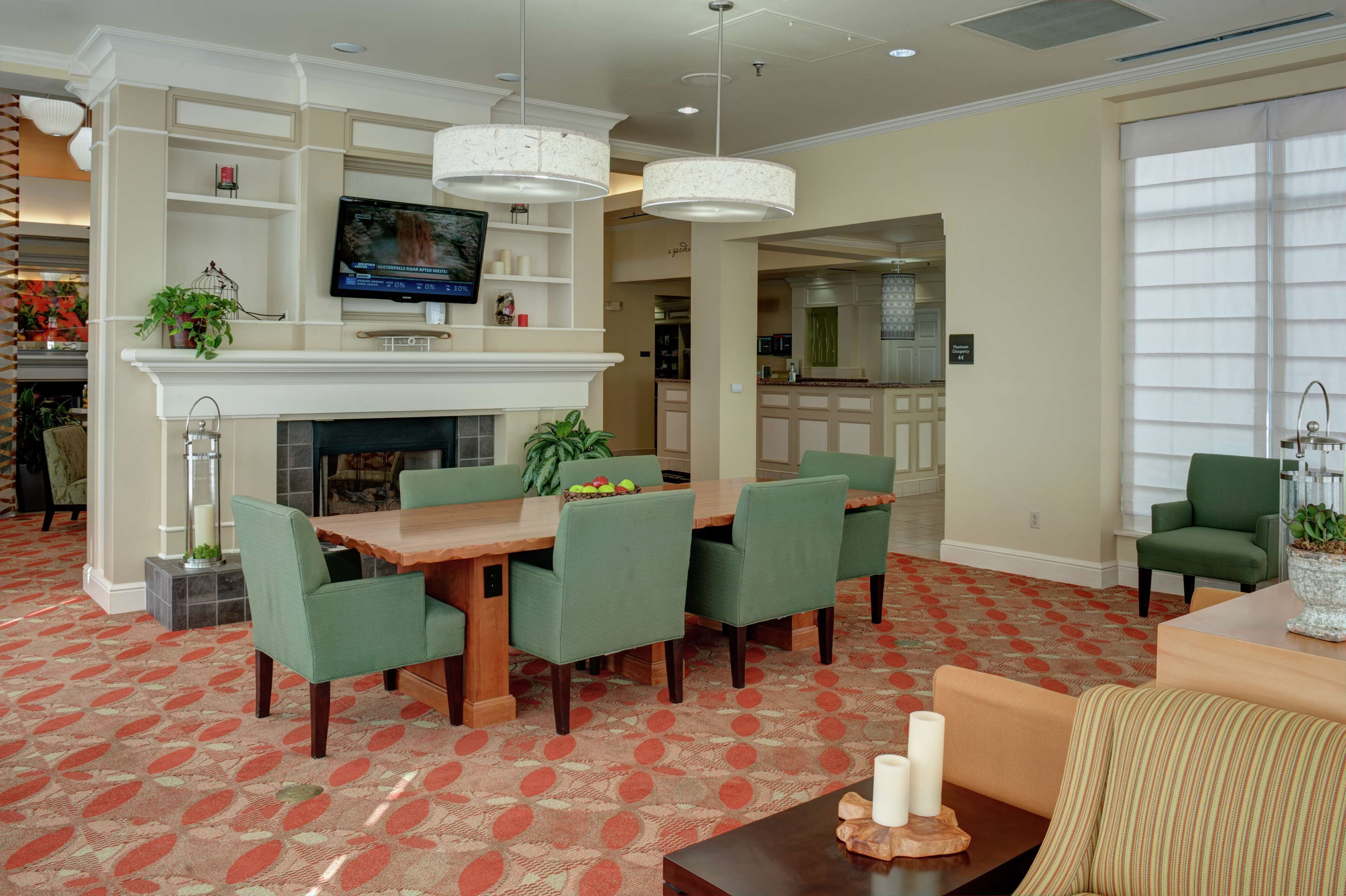 A hotel lobby with a dining area, a fireplace, a television, and a kitchen in the background.