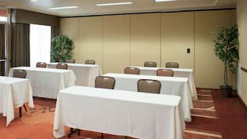 Un salón de conferencias con varias mesas y sillas, una planta grande en maceta y suelo alfombrado.