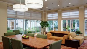 A conference room with a wooden table, green chairs, a sofa, and a coffee table. There are large windows with blinds, a plant, and a view of the outdoors. - Hilton Garden Inn Tulsa Airport (Tulsa)