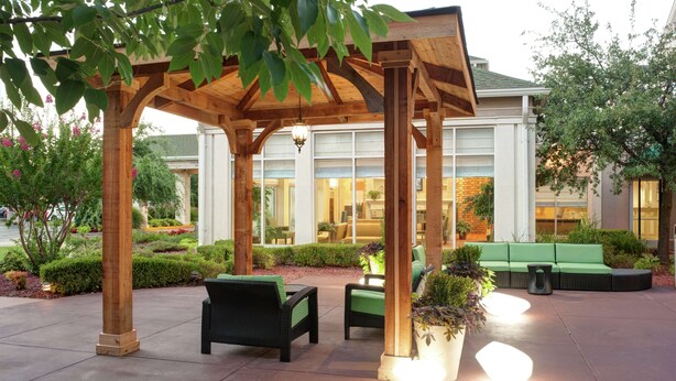 A covered outdoor seating area with wooden beams, green cushioned chairs, and potted plants. - Hilton Garden Inn Tulsa Airport (Tulsa)