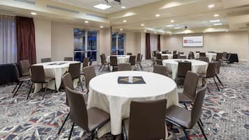 A conference room with multiple round tables and chairs, a projector screen displaying the Hilton Garden Inn logo, and large windows with curtains.