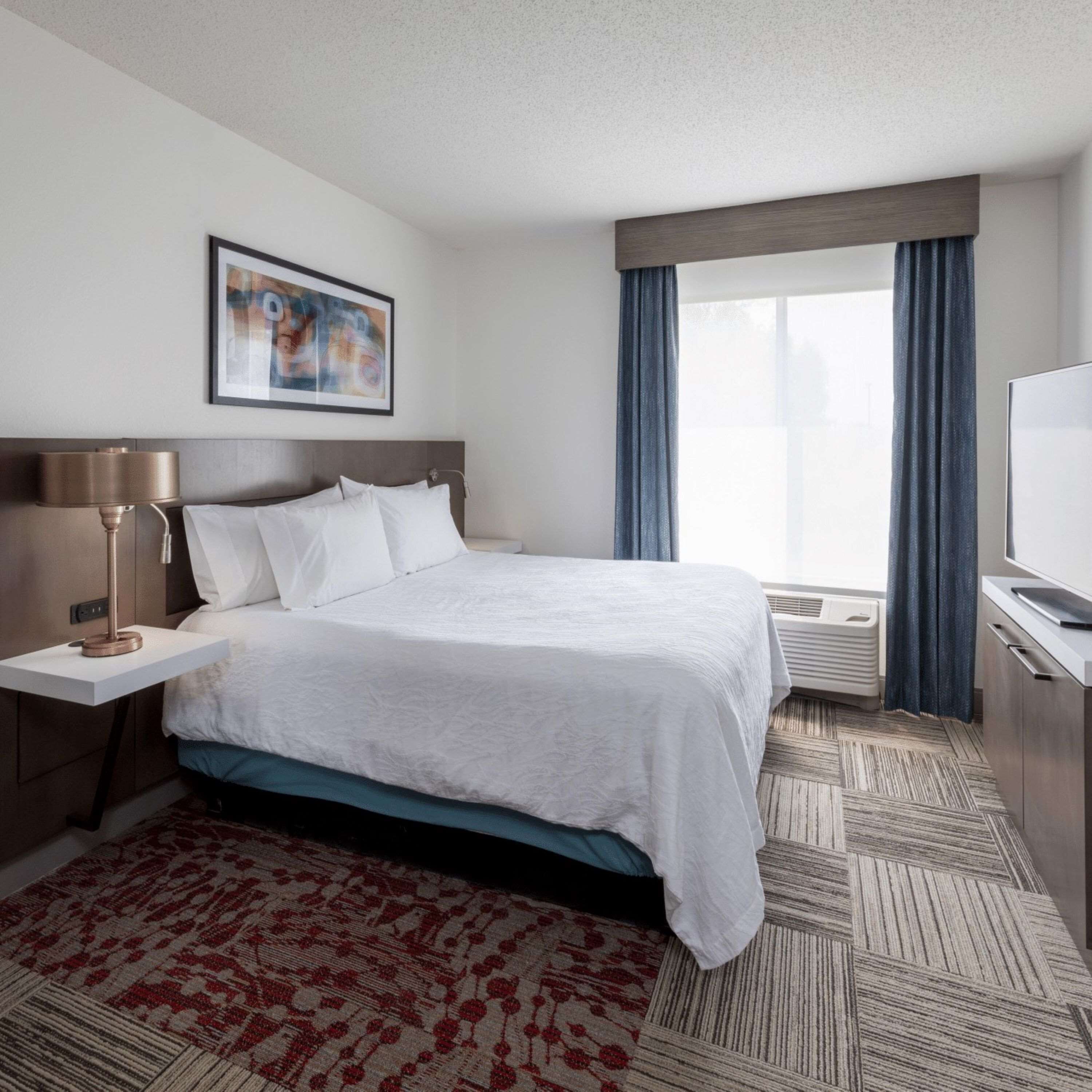 A hotel room with a bed, a bedside table with a lamp, a TV, a window with curtains, and a painting on the wall.