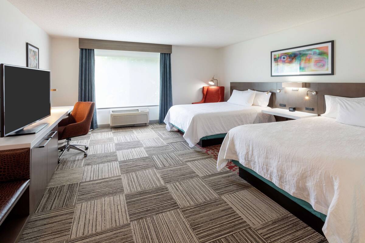 Een hotelkamer met twee bedden, een bureau, een flatscreen televisie, een stoel en een groot raam met gordijnen.