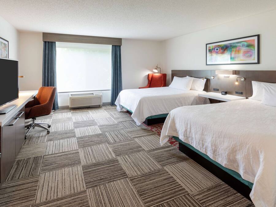 Een hotelkamer met twee bedden, een bureau, een flatscreen televisie, een stoel en een groot raam met gordijnen.